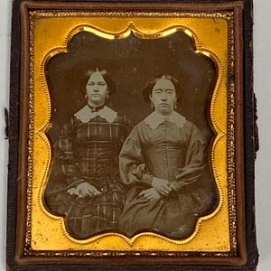 May include: A vintage framed photograph featuring two women in dark dresses with white collars. The image is in a gold-colored, ornate frame with a dark brown border. The women are seated, with their hands clasped in their laps.