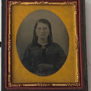 May include: An antique portrait of a woman in a black dress, framed in a rectangular black frame with gold and red accents. The portrait is oval-shaped and features a woman with long hair and earrings.