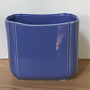 May include: A periwinkle blue ceramic planter with a rectangular shape and rounded corners. The exterior features vertical ribbed detailing along the sides, and the surface has a smooth, glossy finish that reflects light across its curved edges.