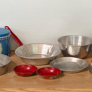 May include: A collection of vintage toy kitchenware including various aluminum pots, pans, and lids with red handles, a blue canister, green plates, a white plastic rack, a small metal kettle, and a silver oval roaster with a matching lid.