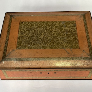May include: A vintage, rectangular, metal box with a hinged lid. The box is decorated with a gold-colored, swirling pattern on a light brown background. The lid has a keyhole and a small metal latch.