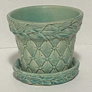 May include: A small, turquoise ceramic planter with a matching saucer. The planter features a quilted diamond pattern and a decorative leaf design around the rim and saucer edge. The pot is designed for small plants.