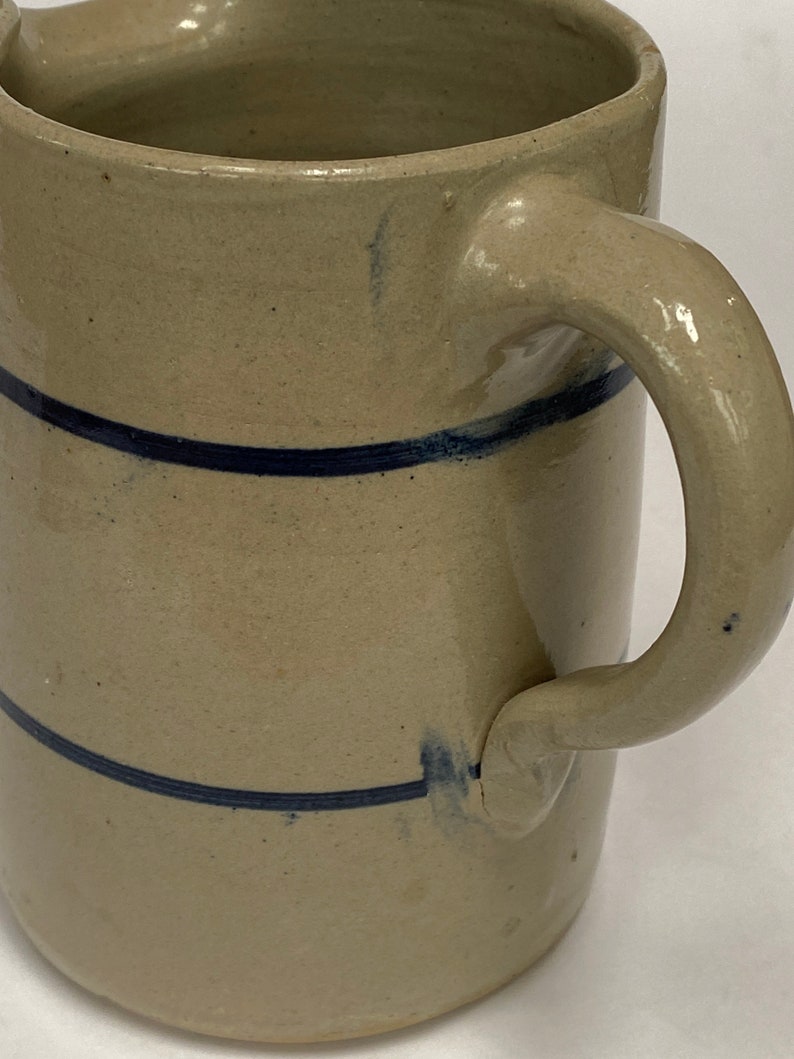 May include: A stoneware pitcher with a blue glaze and two blue stripes around the body. The pitcher has a handle and a wide mouth.