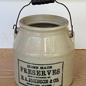 May include: A beige ceramic crock with a wire handle and a black wooden grip. The front features a black rectangular label with the text: HOME MADE PRESERVES MANUFACTURED BY H. A. JOHNSON & CO. BOSTON, MASS. The surface has a visible crackle glaze finish.
