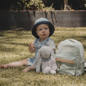 Può includere: Un bambino piccolo con un cappello a quadri blu e bianco è seduto sull'erba con un coniglietto di peluche grigio e uno zaino di tela blu chiaro con il nome "Ollie" ricamato.