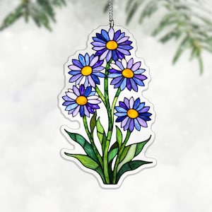 May include: A floral ornament depicting a cluster of blue and purple daisy-like flowers with yellow centres and green leaves. The ornament is suspended by a silver chain. The flowers are in a stained glass style.