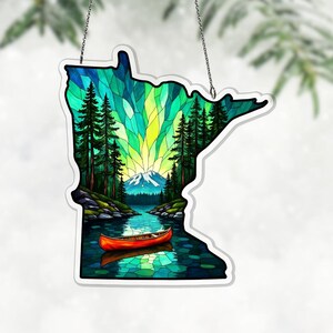 May include: A stained glass-style decoration in the shape of the state of Minnesota, featuring a scenic landscape. The image includes a lake, mountains, trees, and a canoe, with a vibrant color palette of greens, blues, and yellows. It hangs from a silver chain.