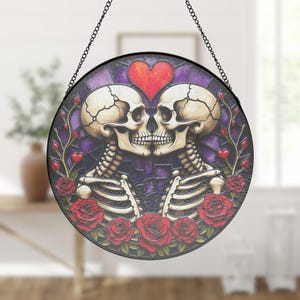 Op de afbeelding: Ronde glas-in-lood kunst met twee kussende skeletten en een rood hart erboven. Het ontwerp omvat rode rozen en een paarse achtergrond. Opgehangen aan een zwarte ketting.