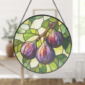 May include: Circular stained glass window art depicting a fig branch with three ripe figs. The figs are purple and red, with green leaves and a yellow background. The window is framed in black and hangs from a chain.