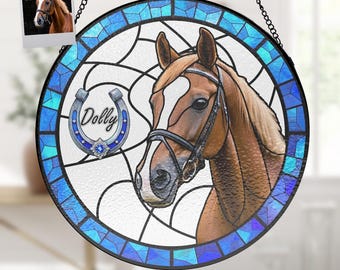 Pode incluir: Janela redonda de vitral com a cabeça de um cavalo com freio. O design inclui uma ferradura azul com o nome "Dolly" e uma gema azul. A borda é um mosaico de peças de vidro azul. Uma pequena foto do cavalo está no canto superior esquerdo.