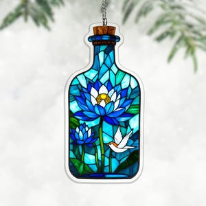 Könnte beinhalten: Ein Glasornament in Flaschenform mit einer blauen Lotusblume mit gelbem Zentrum und einem weißen Vogel. Die Flasche hat einen Korken und wird von einer silbernen Kette gehalten.
