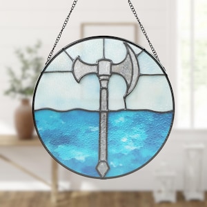 Op de afbeelding: Rond gebrandschilderd glazen raamkunst met een zilveren bijldesign. De bijl staat tegen een achtergrond van blauw en wit glas, dat water en lucht voorstelt. Het kunstwerk is zwart omlijst en hangt aan een ketting.