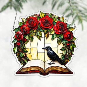 Può includere: Pannello decorativo in vetro colorato con una ghirlanda di rose rosse e foglie verdi. Un corvo nero è appollaiato su un libro aperto. Lo sfondo è un motivo a vetrata giallo chiaro, con una catena d'argento per appenderlo.