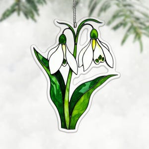 以下が含まれることがあります： 2つのスノードロップの花を特徴とする装飾的な吊り下げオーナメント。花は、緑の葉と茎に対して、緑と黄色のアクセントが付いた白い花びらを持っています。オーナメントはシルバーチェーンで吊り下げられています。