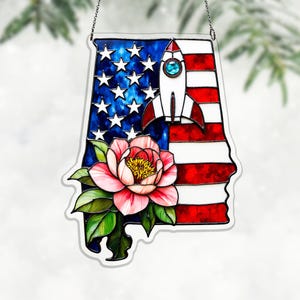 Könnte beinhalten: Glasdekoration in Form des Bundesstaates Alabama, mit der amerikanischen Flagge, einer Rakete und einer rosa Blume. Sie hängt an einer silbernen Kette. Das Design erinnert an die USA.