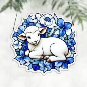 以下が含まれることがあります： 青と白の花の中にいる白い子羊を描いた、装飾用の吊り下げオーナメント。子羊は赤い鼻と茶色のひづめをしています。オーナメントは青い花と葉の縁で囲まれ、吊り下げるための銀色のチェーンが付いています。