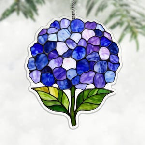 Könnte beinhalten: Ein Glasfenster-Ornament mit einer Hortensienblüte. Die Blüte besteht aus blauen, violetten und weißen Glasteilen. Grüne Blätter und Stiel befinden sich am unteren Rand. Eine silberne Kette ist zum Aufhängen befestigt. Das Ornament ist ein dekorativer Artikel.