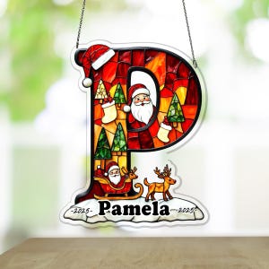 May include: A Christmas-themed hanging decoration in the shape of the letter "P". The decoration features a stained-glass effect with Santa Claus, Christmas trees, and reindeer. The name "Pamela" is displayed, along with the year "2025".