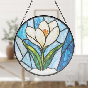 Puede incluir: Vidriera redonda que representa una flor de crocus blanca con un centro naranja y hojas verdes. El fondo es un mosaico de vidrio azul y blanco. Un marco y una cadena de metal negro completan el diseño.
