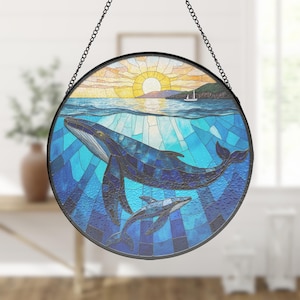 Puede incluir: Un atrapasoles circular de vidrieras que muestra una ballena azul grande y una cría nadando bajo el agua. La escena representa una puesta de sol sobre el océano con un velero distante en el horizonte, en tonos azules, amarillos y blancos.