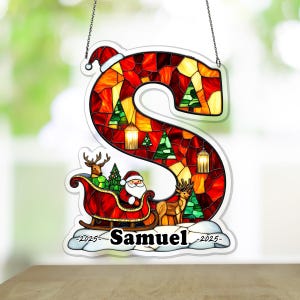 Puede incluir: Adorno navideño de vidriera con forma de letra "S". Presenta a Papá Noel en un trineo con renos, árboles de Navidad y faroles. El nombre "Samuel" se muestra en la parte inferior, con el año 2025.