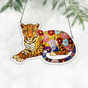 Puede incluir: Un letrero decorativo colgante con un diseño de leopardo con patrones florales coloridos. El leopardo tiene un pelaje manchado naranja y negro, con un contorno blanco. El letrero está suspendido por una cadena plateada.