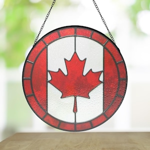 Peut inclure: Vitrail rond représentant le drapeau canadien. Il présente une feuille d'érable rouge sur fond blanc, encadrée de bordures rouges et noires. Une chaîne noire est attachée pour la suspension.