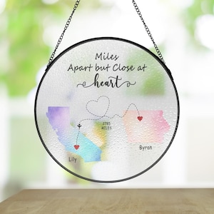 May include: A round glass ornament featuring a watercolour map of two states connected by a dotted line with a heart at the centre. The text "Miles Apart but Close at heart" is written above the map. The states are labelled "Lily" and "Byran" with the distance between them labelled "2785 Miles".
