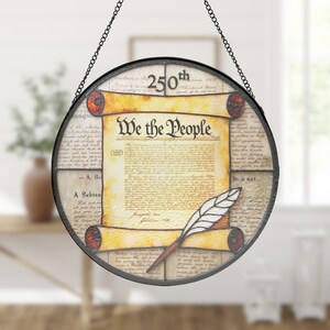 May include: A round, stained-glass window hanging with a black metal frame and chain. The design features a parchment with the words "We the People" and a quill pen. The background includes printed text and the number "250th".
