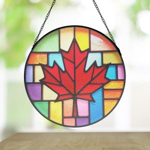 May include: A round stained glass window with a black metal frame and hanging chain. The window features a red maple leaf design in the center, surrounded by colorful geometric shapes in shades of yellow, green, blue, and purple.