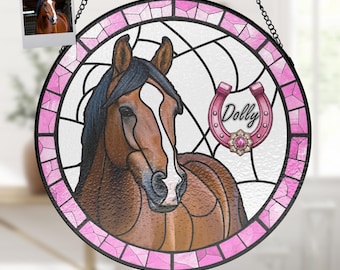 Pode incluir: Uma janela de vitral pendurada com um design rosa e branco. O centro apresenta um cavalo castanho com uma crina preta e uma ferradura rosa com o nome "Dolly" nela.