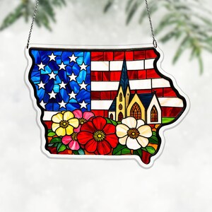 May include: Stained glass Iowa state outline with an American flag design, a church, and colorful flowers. The piece is framed with a white border and hangs from a silver chain. The flag features red and white stripes and blue with white stars.
