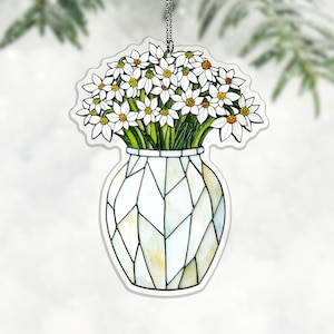 Könnte beinhalten: Dekoratives Ornament mit einem Strauß weißer Blumen mit gelben Zentren in einer weißen Vase. Die Vase hat ein geometrisches Design. Das Ornament hängt an einer silbernen Kette. Die Blumen haben grüne Stiele und Blätter.