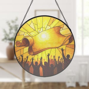 May include: Round stained glass window with a black metal frame and chain. The design features a golden scroll and a crowd of people with raised arms against a yellow background. The glass is textured and the image is reminiscent of a religious scene.