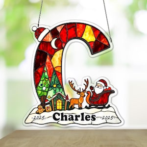 Puede incluir: Adorno navideño de vidriera con forma de letra "C". El adorno presenta a Papá Noel en un trineo, un reno, un árbol de Navidad y el nombre "Charles". El adorno es rojo, amarillo y verde, con el año 2025.
