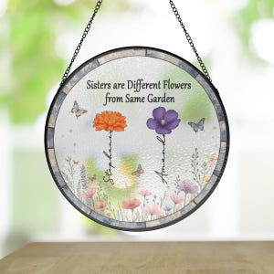May include: Round stained glass suncatcher with a floral design and the quote "Sisters are different flowers from the same garden". The names Stephanie and Amanda are written vertically next to the flowers.