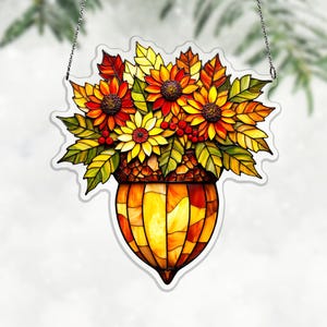 Puede incluir: Decoración de ventana de vidriera con un ramo de girasoles y hojas de otoño en un jarrón en forma de bellota. Los girasoles son amarillos y rojos, con hojas verdes y bayas rojas. El jarrón es un mosaico de vidrio naranja y amarillo.