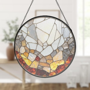May include: A round stained glass window with a black metal frame and chain for hanging. The glass features a mosaic design with shades of white, gray, brown, yellow, and red. The window is a decorative home accent.
