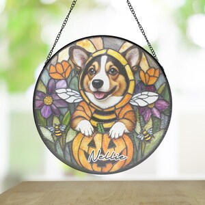 May include: Round stained glass window art featuring a corgi dressed as a bee, smiling and sitting on a pumpkin. The artwork includes flowers, bees, and the name "Nellie" on the pumpkin. The window is framed in black and hangs from a chain.