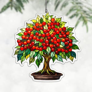 Op de afbeelding: Een decoratief ornament met een bonsai-boom vol helderrode kersen. De bladeren zijn een mix van groen en geel en de stam is bruin. De boom staat in een bruine pot en hangt aan een zilveren ketting.