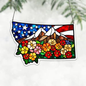 Puede incluir: Vidriera con forma de Montana, con montañas, flores y la bandera estadounidense. La obra utiliza colores vibrantes como azul, rojo, blanco, amarillo y naranja. Cuelga de una cadena plateada. Mide unos 25 cm de ancho.