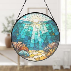 Puede incluir: Un atrapasoles circular de vidrieras que muestra una escena submarina con un sol brillante en la parte superior, rayos de luz filtrándose a través del agua azul y turquesa, un fondo arenoso y coloridos arrecifes de coral con pequeños peces.