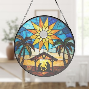 May include: Round stained glass window art with a black metal frame and chain. The design features a vibrant sun, palm trees, and a nativity scene. The colors include blue, yellow, orange, and brown. This decorative piece is suitable for indoor or outdoor display.
