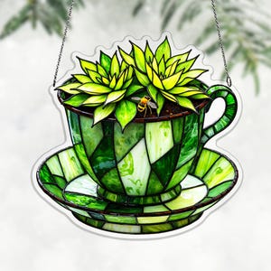 May include: A stained glass teacup and saucer planter with a handle, filled with green succulents and a small bee. The teacup and saucer are made of green glass pieces, and the succulents have bright green leaves. The planter hangs from a silver chain.
