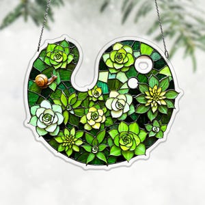 May include: A stained glass succulent design in shades of green, with a small snail. The artwork is in a unique shape and is suspended by a chain. The piece features various succulent flowers and leaves.