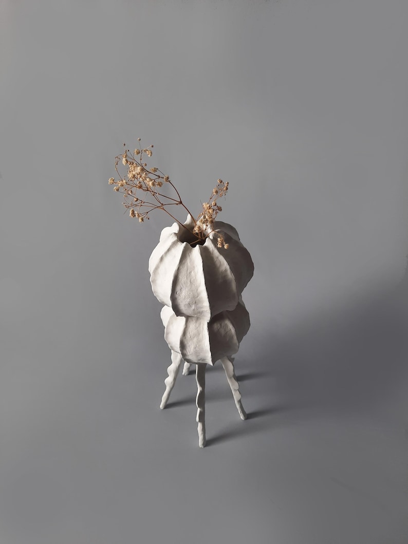 May include: White ceramic vase with a unique, organic shape and three legs. The vase is filled with dried flowers.