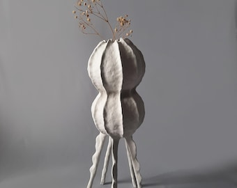 Könnte beinhalten: Weiße Keramikvase mit einer strukturierten, organischen Form. Die Vase hat einen breiten, runden Boden und einen hohen, schlanken Hals. Sie wird von vier langen, dünnen Beinen getragen. Die Vase ist mit getrockneten Blumen gefüllt.