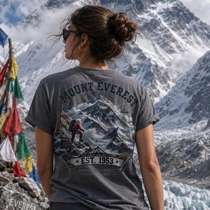 Puede incluir: Una camiseta gris carbón de manga corta con un estampado gráfico del Monte Everest, un escalador y banderas de oración. El texto dice MOUNT EVEREST SUMMIT, FIRST ASCENT y EST. 1953. Se combina con pantalones cargo de color gris oscuro.