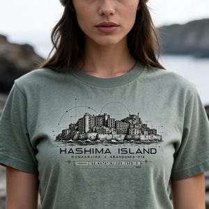 May include: A sage green short-sleeve t-shirt featuring a black ink-style graphic of Hashima Island. The print includes the text HASHIMA ISLAND, GUNKANJIMA, ABANDONED 1974, and the coordinates 22.6376 N, 129.7385 E with technical line drawings.