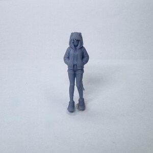 May include: A small, gray figurine of a person wearing a hooded jacket, shorts, and sneakers. The hood has cat-like ears. The figure is standing with hands in pockets, suggesting a casual pose. The overall aesthetic is minimalist.